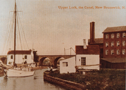 This colorized 1908 postcard shows a sailboat departing Lock No. 13, known as the “Deep Lock” in the City of New Brunswick.   Although not the deepest lock on the Delaware and Raritan Canal (Lock No. 4 south of Lalor Street in Trenton was deeper after it was combined with Lock No. 5 in 1853) Lock No. 13 did raise and lower vessels 12.2 feet.  The locktender’s house can be seen behind the boat, while the Johnson & Johnson factory buildings are visible to the right.  Lock No. 13 was filled in order to facilitate the construction of N.J. Highway Route No. 18 through New Brunswick.